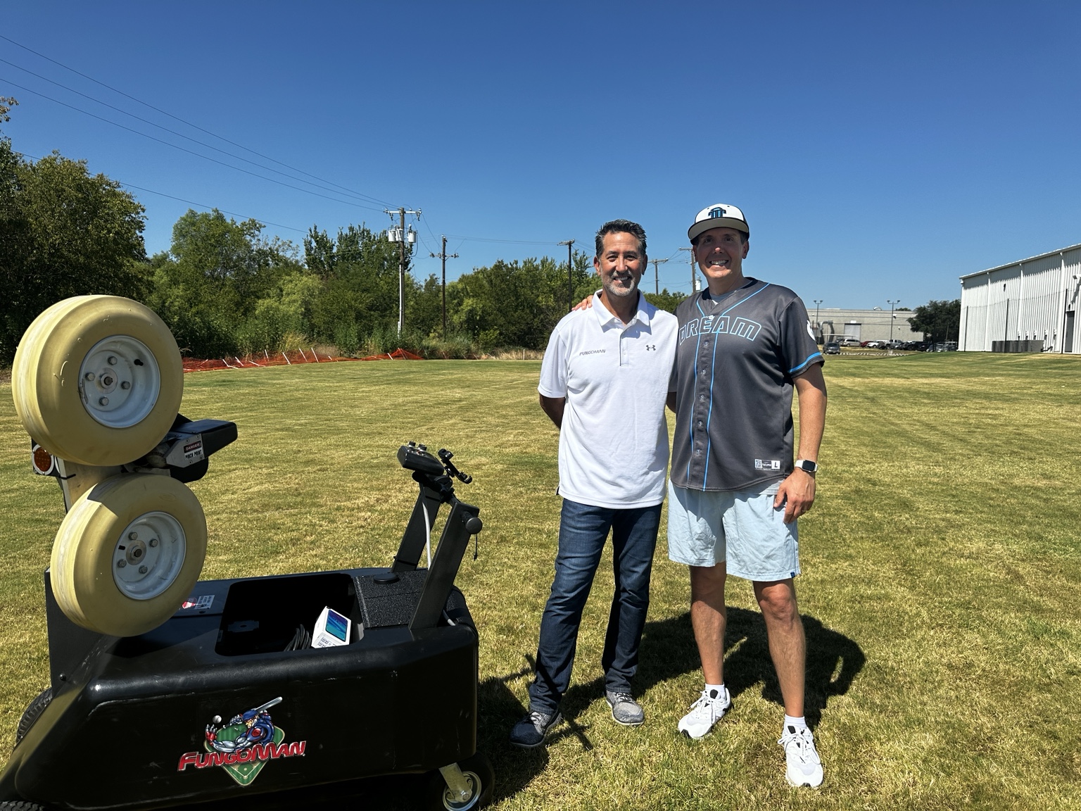 Patrick with another adult beside a pitching machine on a grass field